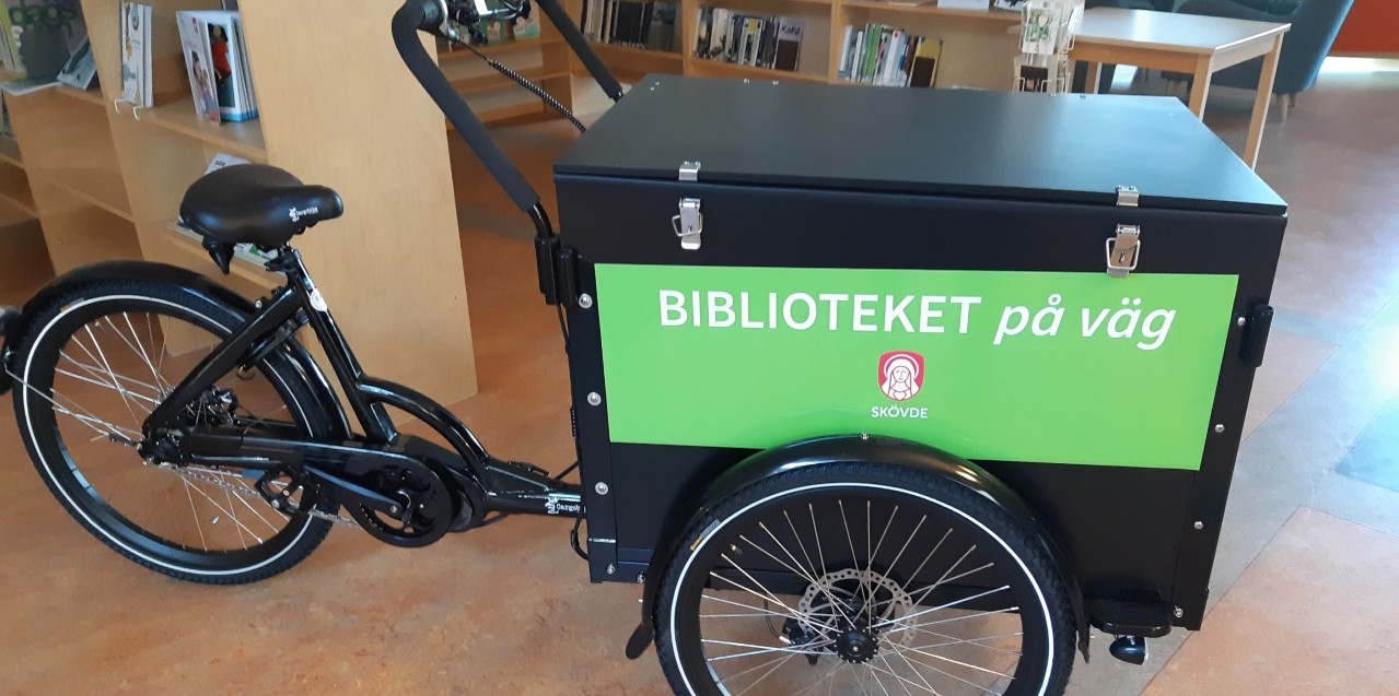 En svart cykel med låda och grön skylt med texten Biblioteket på väg